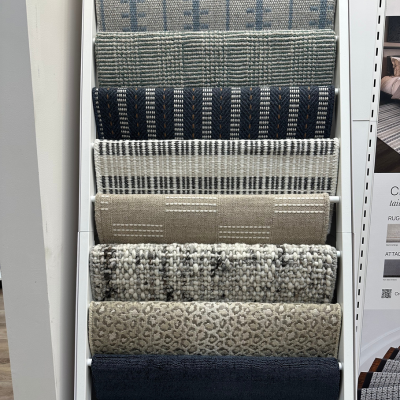 Vertical rack with rolled-up rug samples in diverse patterns and textures, including stripes, animal prints, and herringbone, alongside a partial display board with a staircase image.